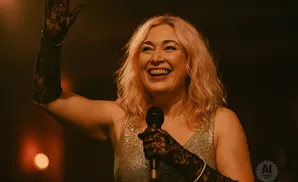 A smiling woman with blonde hair and a sparkly dress holds a microphone and raises her left hand.