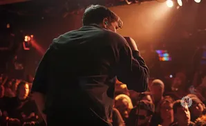 A singer on stage at a concert, facing away from the camera, with an audience visible in the background.