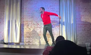 A man in a red sweater and dark pants stands on stage, smiling, with a brick wall and curtains behind him.