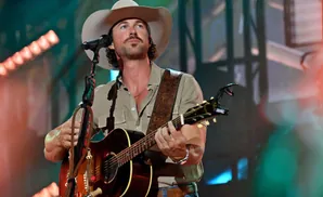 A man wearing a cowboy hat and a light brown shirt plays an acoustic guitar on stage.