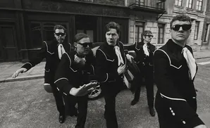 Five men in black suits and sunglasses stand on a street.