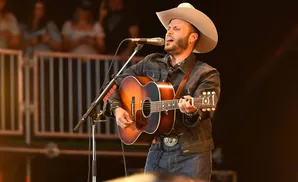 A man in a cowboy hat sings into a microphone while playing an acoustic guitar on stage.