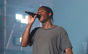 A Black man with braids sings into a microphone on stage, wearing a grey t-shirt and jewelry.