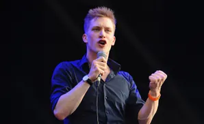 A man with spiky blonde hair, wearing a dark shirt, speaks into a microphone while clenching his fist.