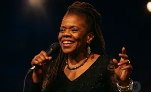 A Black woman with dreadlocks sings into a microphone, smiling broadly and wearing a sparkly black dress.