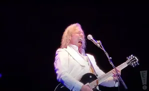 A male singer in a white suit plays a black electric guitar on stage.
