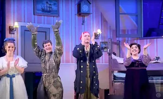 Four actors on a stage, two men and two women, clap with raised hands in front of a room with striped wallpaper.