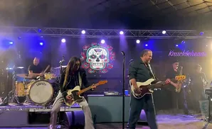 A rock band performs on stage with a colorful skull backdrop and neon 