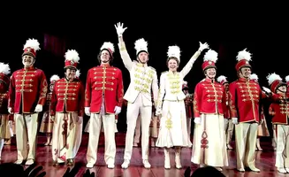 A cast in red and white marching band uniforms on stage, some waving.