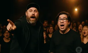 Two men in black shirts and a choir sing with mouths open, one pointing forward.