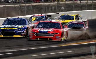 NASCAR race cars in a close pack, kicking up dust on the track.