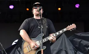 Musician wearing sunglasses and a black t-shirt plays a guitar on stage, with orange-patterned shorts visible.