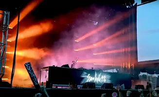 A DJ performs on stage at a concert, bathed in orange and pink stage lights and smoke.