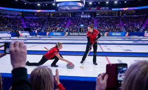 Two women in red curling uniforms play on an ice rink with a crowd watching in the background.