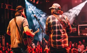 Two men play guitar on stage at a concert, facing away from the camera.