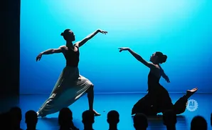 Two dancers perform on a stage with a blue backdrop; one in a light skirt, the other in dark pants.