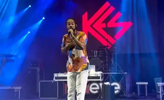 Man in a colorful shirt sings into a microphone on a stage with blue lights and a large pink logo.