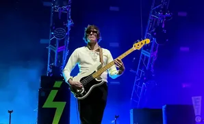 A male bassist plays a white Fender Precision bass on a stage illuminated by blue lights.