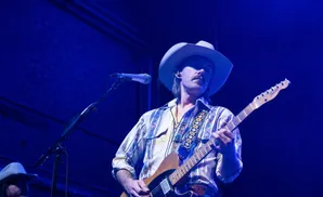 A man wearing a cowboy hat and a patterned shirt plays an electric guitar under blue stage lights.