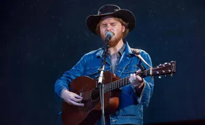 Musician in a cowboy hat and denim jacket plays an acoustic guitar and sings into a microphone.