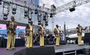 A group of male singers in matching patterned jackets and yellow pants perform on an outdoor stage with lighting rigs above.