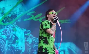 Man in a green and black tie-dye shirt and neon green glasses sings into a pink microphone on stage.