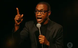 A Black man with glasses holds a microphone and points his finger upwards, speaking animatedly.