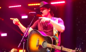 A country singer in a cowboy hat points while playing an acoustic guitar on a stage lit with pink and red lights.