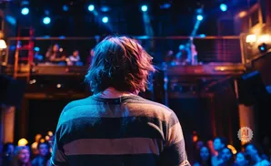 Person with wavy brown hair facing away from camera, wearing a striped sweater, on stage with a crowd and blue lights.