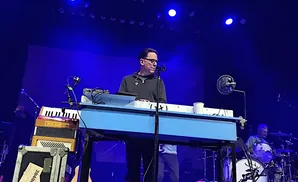 A man in glasses plays a keyboard on stage, with a drummer in the background.