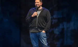 A man in a black hoodie and jeans smiles while holding a microphone on stage.