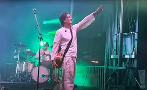 A male musician in a white shirt and pants plays guitar on stage, pointing his finger to the right, with a drummer in the background.