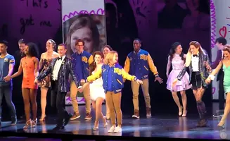 A musical theater cast, some in letterman jackets, takes a bow on stage.