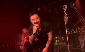 A male singer with tattoos and spiky hair sings into a microphone on stage, with a guitarist behind him.