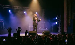 A man in a suit speaks into a microphone on stage, with a projection screen behind him and an audience in the foreground.