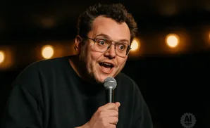 A man with glasses and an open mouth holds a microphone, speaking to an audience with blurred lights behind him.