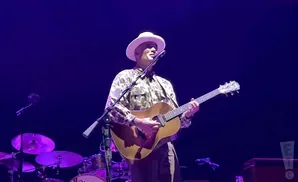 A man in a white hat and camouflage shirt plays an acoustic guitar on stage.
