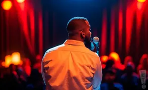 A man stands with his back to the camera, speaking into a microphone on a stage with red lights and a blurred audience.