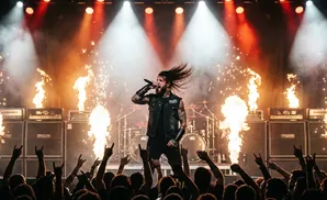 A male singer with long hair and a vest performs on stage with flames and fireworks, while a crowd holds up rock on sign.