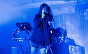 Singer in a dark shirt and denim shorts, holding a microphone with a blue stage light.