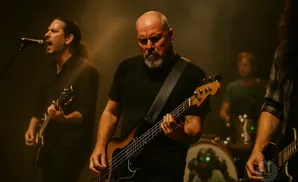 A bald man with a goatee plays a bass guitar on stage with a band.