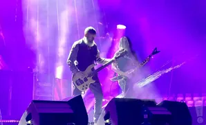 Two guitarists perform on a purple-lit stage, one with a beard playing a light-colored bass guitar.