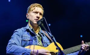 Musician with blonde hair plays an acoustic guitar and sings into a microphone while wearing a denim jacket.
