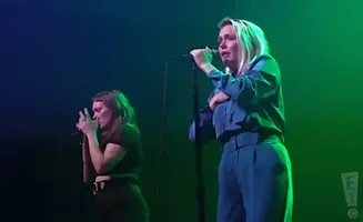 Two women sing into microphones on a dimly lit stage with green and blue lights.