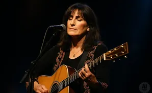 Woman with eyes closed plays an acoustic guitar on stage, with a microphone in front of her.