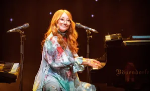 A redheaded woman in glasses plays a Bösendorfer piano on stage.