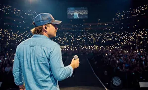 A man in a baseball cap and denim shirt holds a microphone on stage, facing a cheering crowd with their phones lit up.