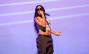 A woman in sunglasses and a crop top sings into a microphone on a purple stage.
