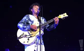 Musician with curly hair plays a white electric guitar on stage, wearing a tie-dye shirt and white pants.