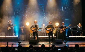 A four-man band performs on stage with blue spotlights, two singers with guitars, a bassist, and a drummer.